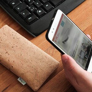 Puede incluir: Una funda de tel&eacute;fono de corcho marr&oacute;n con la palabra "Squips" impresa en ella. Una persona sostiene un tel&eacute;fono inteligente blanco con una funda negra. La pantalla del tel&eacute;fono muestra un sitio web con el texto "K-U" y una imagen de unas gafas.