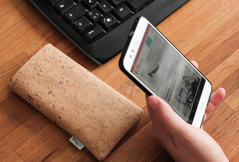 Puede incluir: Una funda de tel&eacute;fono de corcho marr&oacute;n con la palabra "Squips" impresa en ella. Una persona sostiene un tel&eacute;fono inteligente blanco con una funda negra. La pantalla del tel&eacute;fono muestra un sitio web con el texto "K-U" y una imagen de unas gafas.