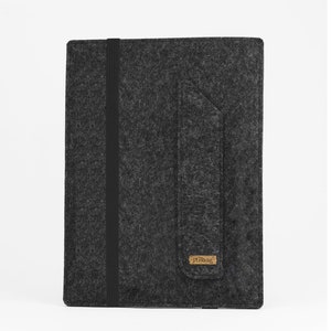 May include: A dark gray felt sleeve for a tablet or notebook, featuring a vertical black elastic band closure and a side pocket flap. A small rectangular brown label with the text stilbag is attached to the bottom corner of the pocket.