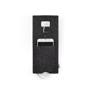 Op de afbeelding: Een grijze telefoonoplader van vilt met een witte telefoon erin en een witte oplader die aan de muur is bevestigd.