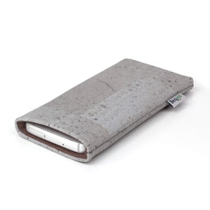 Puede incluir: Una funda para tel&eacute;fono gris hecha de corcho con un sutil patr&oacute;n moteado. La funda tiene un ribete de cuero marr&oacute;n y un peque&ntilde;o logotipo "shibumi" verde y blanco en el lateral.