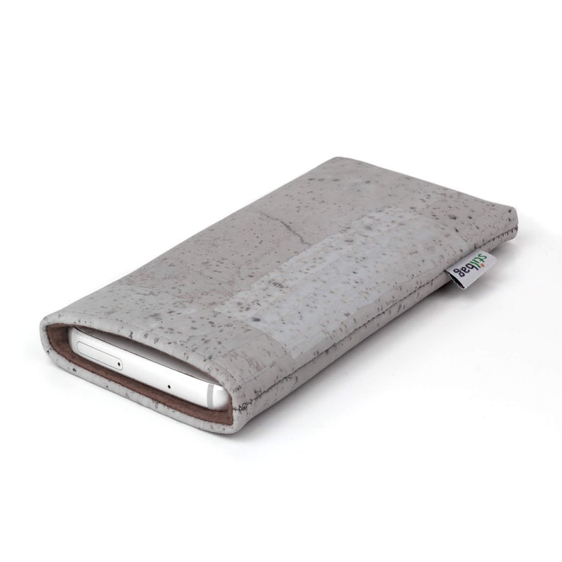 Puede incluir: Una funda para tel&eacute;fono gris hecha de corcho con un sutil patr&oacute;n moteado. La funda tiene un ribete de cuero marr&oacute;n y un peque&ntilde;o logotipo "shibumi" verde y blanco en el lateral.