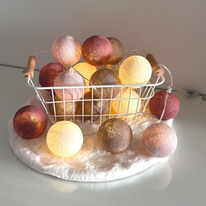 Könnte beinhalten: Ein weißer Drahtkorb gefüllt mit einer Lichterkette aus Baumwollkugeln. Die Lichter sind in verschiedenen Schattierungen von Rosa, Beige und Braun. Die Lichter liegen auf einer weißen, flauschigen Oberfläche.