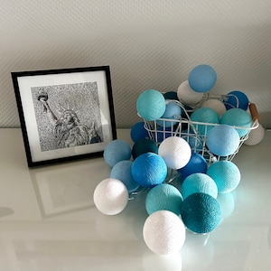 Könnte beinhalten: Ein weißer Drahtkorb gefüllt mit blauen und weißen Baumwollkugeln. Die Lichter sind kreisförmig angeordnet und leuchten. Der Korb steht auf einer weißen Oberfläche.