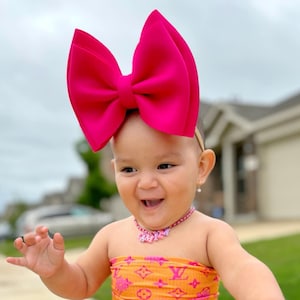 May include: A smiling baby wearing a bright pink bow headband, a matching pink necklace, and a strapless orange dress with a pink pattern. The outfit is completed with pink fluffy leg warmers and white sneakers. The baby is standing on a sidewalk.