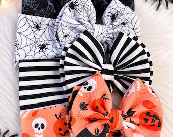 May include: A set of four hair bows attached to headbands featuring Halloween patterns. The designs include a black spiderweb print, a white spiderweb print, black and white horizontal stripes, and an orange fabric with skulls, bats, and pumpkins.