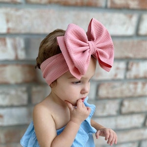 May include: A pink fabric headband with a large, oversized bow. The headband is worn by a young child with brown hair.