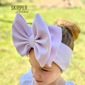 May include: A large, lavender-colored bow headband. The bow is made of a textured fabric and is tied in a large, floppy knot. The headband is worn by a young girl with brown hair.