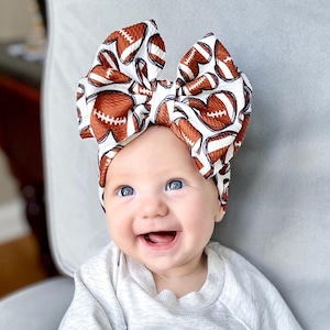 May include: A baby wearing a white headband with a brown and white football pattern. The baby is smiling and looking to the right.