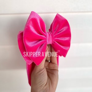 May include: A vibrant pink velvet bow with the text "SKIPPER AVENUE" is held up against a white background. The bow is large and fluffy, with a soft, luxurious texture. The bow is a bright, eye-catching accessory.