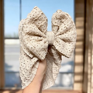 May include: A beige, oversized bow headband. The headband is made of a textured fabric with small, evenly spaced holes. The bow is large and prominent, with a knotted center. The background is blurred, with a window and curtains visible.