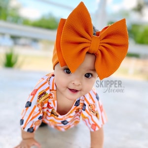 May include: A baby wearing an orange bow headband and a short-sleeved shirt with a burger and fries pattern. The shirt has orange and white stripes. The text "SKIPPER Avenue" is visible.