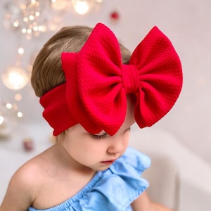 May include: A red fabric headband with a large, oversized bow. The headband is worn by a young child with blonde hair.
