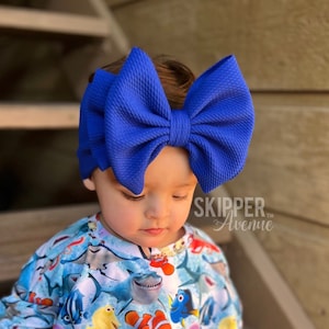 May include: A toddler wearing a blue and white long-sleeved shirt with a print of various sea creatures, including Nemo, Dory, and sharks. The toddler is wearing a large, bright blue bow headband. The toddler is sitting on a wooden step with the text "SKIPPER Avenue" visible in the background.