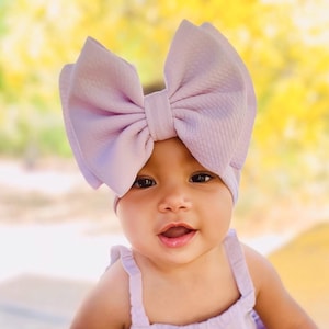 May include: A baby girl wearing a lavender headband with a large bow and a matching lavender dress. The baby is sitting on a wooden surface.