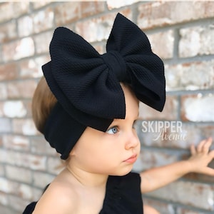 May include: A black headband featuring a large, textured fabric bow. The material has a subtle ribbed pattern. The text Skipper Avenue is visible in white on the right side. The accessory is designed to be worn around the head as a hair piece.