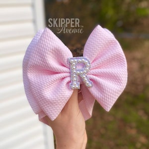 May include: A pink bow with a pearl-encrusted silver letter "R" in the centre. The bow is held in a hand against a white background. The text "SKIPPER Avenue™" is visible above the bow.