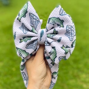 May include: White baby headband with a large bow featuring a pattern of green fish and fishing nets. The bow is tied in the center and has a textured appearance. The background is a blurred green lawn.