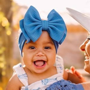 May include: A smiling baby wearing a large blue bow headband and a white and blue patterned outfit. The baby is being held by someone wearing a blue top with lace detailing.