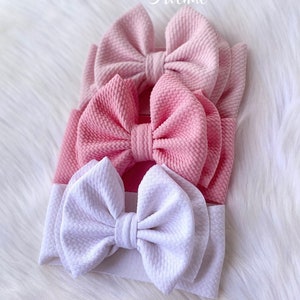 May include: Three pink and white fabric headbands with large bows. The headbands are stacked on top of each other. The top headband is pink, the middle headband is pink, and the bottom headband is white. The headbands are made of a soft, stretchy fabric. The bows are large and fluffy. The headbands are perfect for adding a touch of style to any outfit.