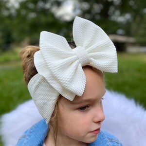 May include: A white, textured, oversized bow headband. The headband is worn by a child with blonde hair and a blue denim jacket.