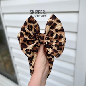 May include: A large brown and black leopard print bow. The bow is tied in a knot and is being held by a hand.