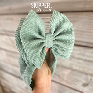 May include: A large, sage green bow headband with a textured fabric. The bow is centered and has a knotted design. The text "SKIPPER Avenue" is printed in white on the top left of the image. The headband is held by a hand.