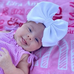 May include: A baby wearing a light purple dress and a large white bow headband. The baby is lying on a pink blanket with text. The dress has tie details on the shoulders. The baby is smiling.
