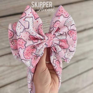 May include: A pink and white patterned bow with the text "SKIPPER Avenue". The bow is held up against a wooden background. The bow has a repeating pattern of pink shapes with red and white accents.