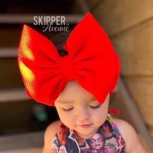 Op de afbeelding: Een rood strik hoofdband op het hoofd van een kind. Het hoofdband is gemaakt van een zachte, textuurrijke stof. Het kind draagt een kleurrijk shirt met een Stranger Things-thema.