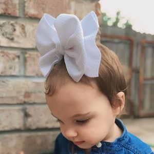 May include: A large white bow hair accessory atop a child's head. The bow is made of a textured fabric and is centered on the head. The child is wearing a blue denim shirt and is looking down. A brick wall is in the background.