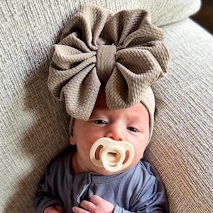 May include: A beige textured fabric headband featuring a large, oversized bow detail worn on the head, paired with a cream-colored silicone pacifier and a long-sleeved dark gray garment. The headband has a gathered, pleated appearance.