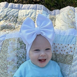 May include: A smiling baby wearing a light blue long-sleeved shirt with a decorative lace trim and a large white bow headband. The baby is lying on a patchwork quilt with floral and geometric patterns.