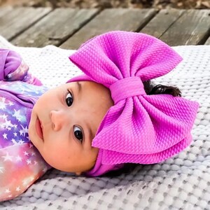 May include: A baby wearing a purple bow headband and a matching onesie. The onesie features a colorful galaxy print with stars. The baby is lying on a gray blanket.