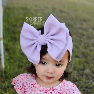May include: A lavender bow headband with a large, fluffy bow. The headband is made of soft, stretchy fabric and is perfect for little girls.