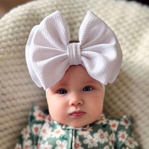 May include: A baby wearing a white bow headband and a green floral print onesie. The baby is lying on a cream-colored, textured surface. The onesie has a zipper closure and ruffle details.