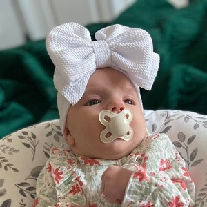 May include: A baby wearing a white bow headband and a floral print onesie. The baby is using a pacifier and is seated in a patterned baby lounger. The background includes a green blanket.
