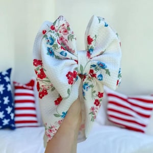 May include: A large white fabric bow with a floral pattern in red, blue, and green. The bow is held up, showcasing its textured surface and vibrant colors. The bow is a hair accessory.