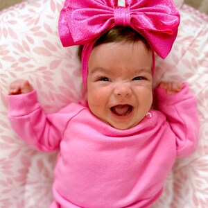Puede incluir: Un beb&eacute; con un atuendo rosa y una gran diadema de lazo rosa. El beb&eacute; sonr&iacute;e y tiene una expresi&oacute;n feliz.