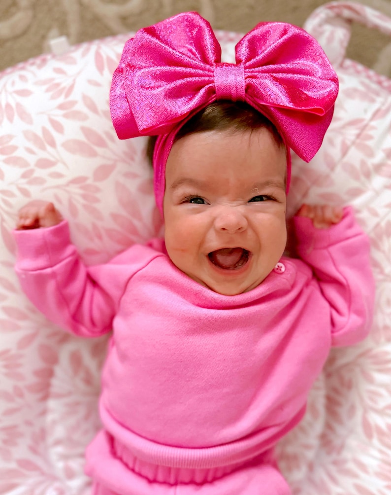 Puede incluir: Un beb&eacute; con un atuendo rosa y una gran diadema de lazo rosa. El beb&eacute; sonr&iacute;e y tiene una expresi&oacute;n feliz.