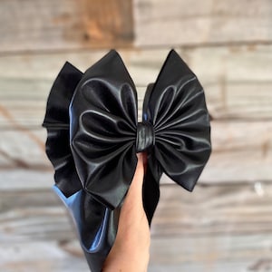 May include: A large black faux leather bow with a ruffled edge. The bow is tied in a knot and has a shiny finish.