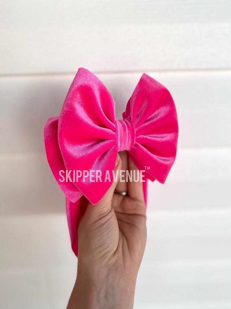 May include: A vibrant pink velvet bow with the text "SKIPPER AVENUE" is held up against a white background. The bow is large and fluffy, with a soft, luxurious texture. The bow is a bright, eye-catching accessory.