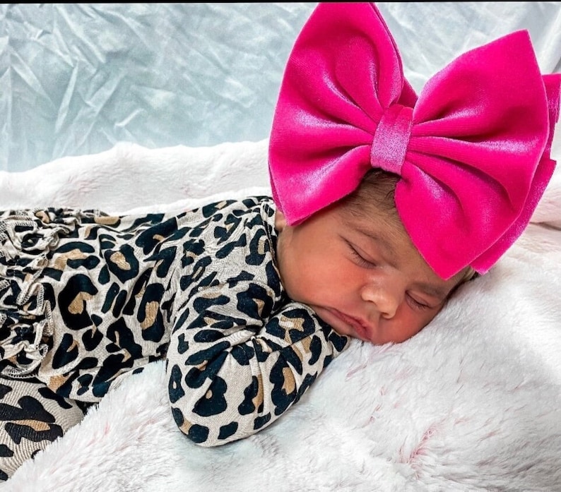 May include: A baby wearing a brown and black leopard print sleeper and a pink velvet bow headband.