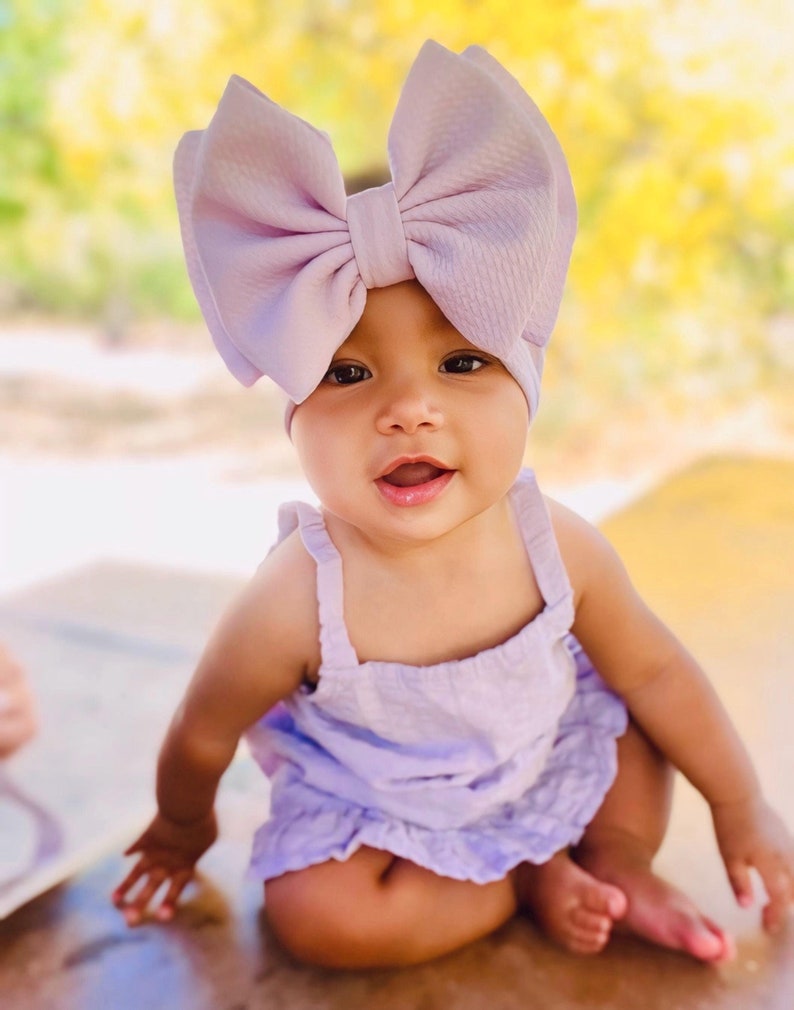 May include: A baby girl wearing a lavender headband with a large bow and a matching lavender dress. The baby is sitting on a wooden surface.