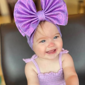 Puede incluir: Un beb&eacute; con una diadema morada con un gran lazo brillante y un vestido morado sin mangas a juego.