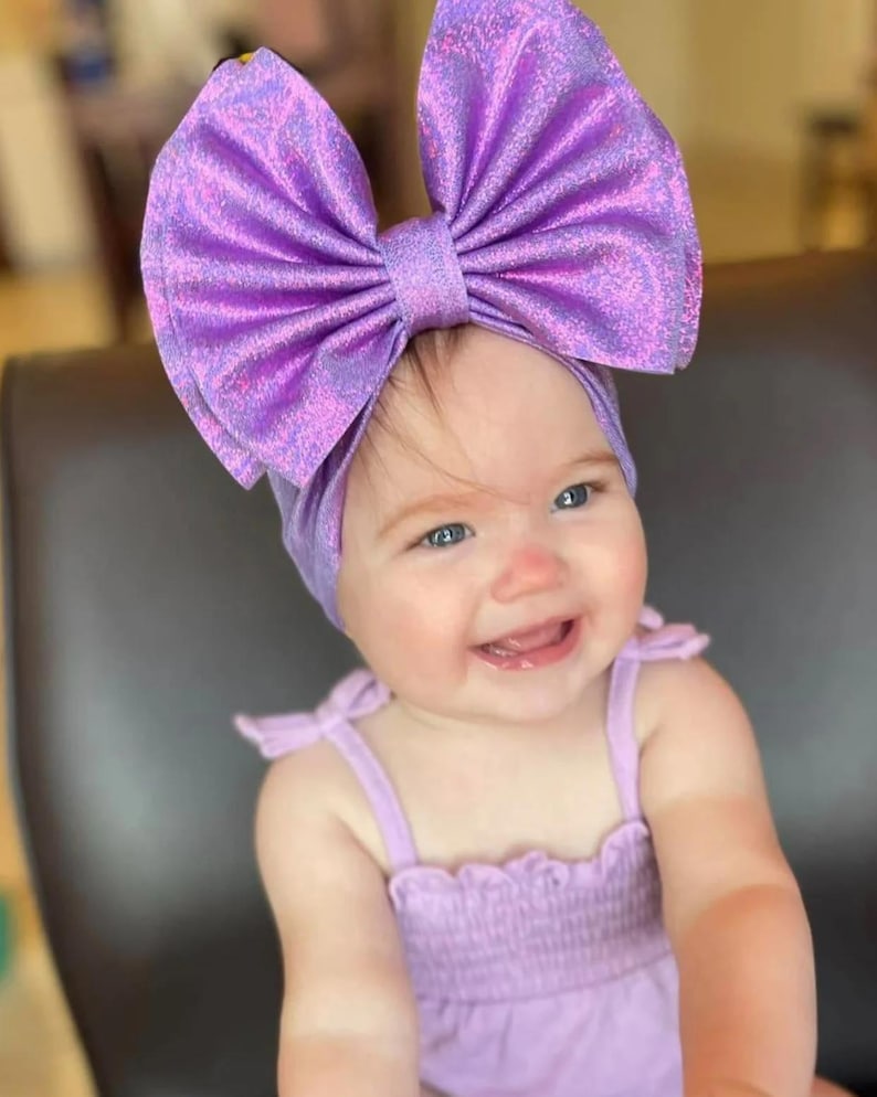 Puede incluir: Un beb&eacute; con una diadema morada con un gran lazo brillante y un vestido morado sin mangas a juego.