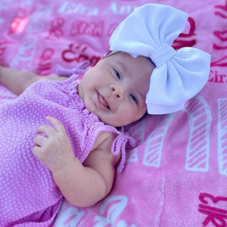 May include: A baby wearing a light purple dress and a large white bow headband. The baby is lying on a pink blanket with text. The dress has tie details on the shoulders. The baby is smiling.