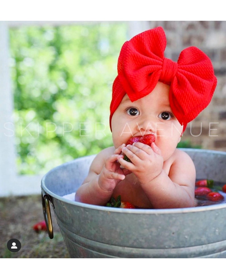 May include: A baby wearing a red bow headband sits in a metal tub filled with water and strawberries. The baby is holding a strawberry in their hand and is looking at the camera.