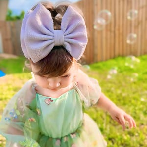 May include: A young girl wearing a light green tulle dress with floral embroidery and a large lavender bow headband. She is standing in a grassy area with a wooden fence in the background.