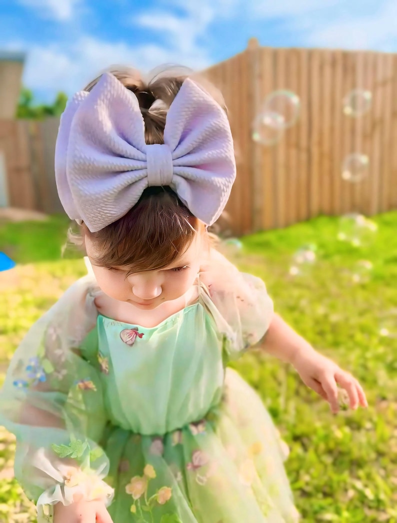 May include: A young girl wearing a light green tulle dress with floral embroidery and a large lavender bow headband. She is standing in a grassy area with a wooden fence in the background.